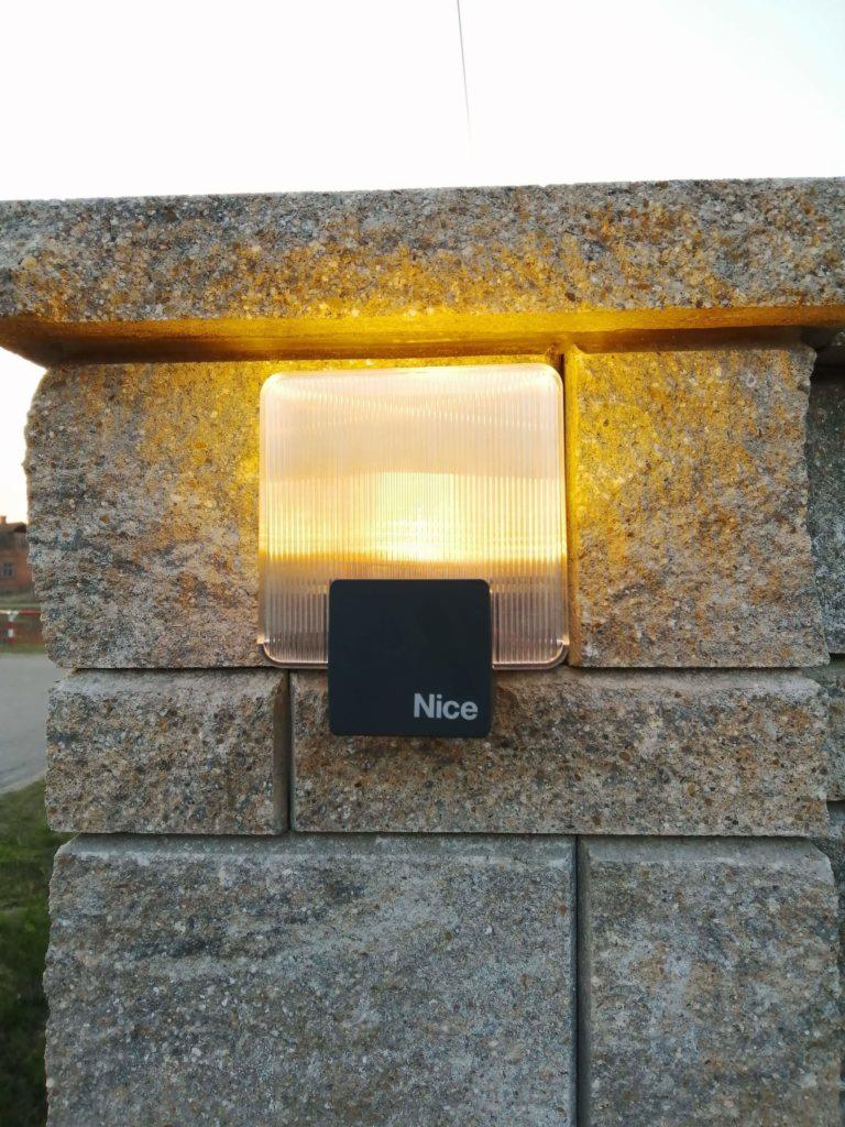 Kwadratowa lampa zewnętrzna z logo 'Nice' zamontowana na kamiennym słupku ogrodzenia, oświetlona ciepłym światłem.
