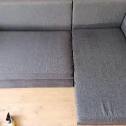 Fragment szarej, modułowej sofy narożnej z widocznym ssawką odkurzacza piorącego na drewnianej podłodze, sugerujący czyszczenie tapicerki.