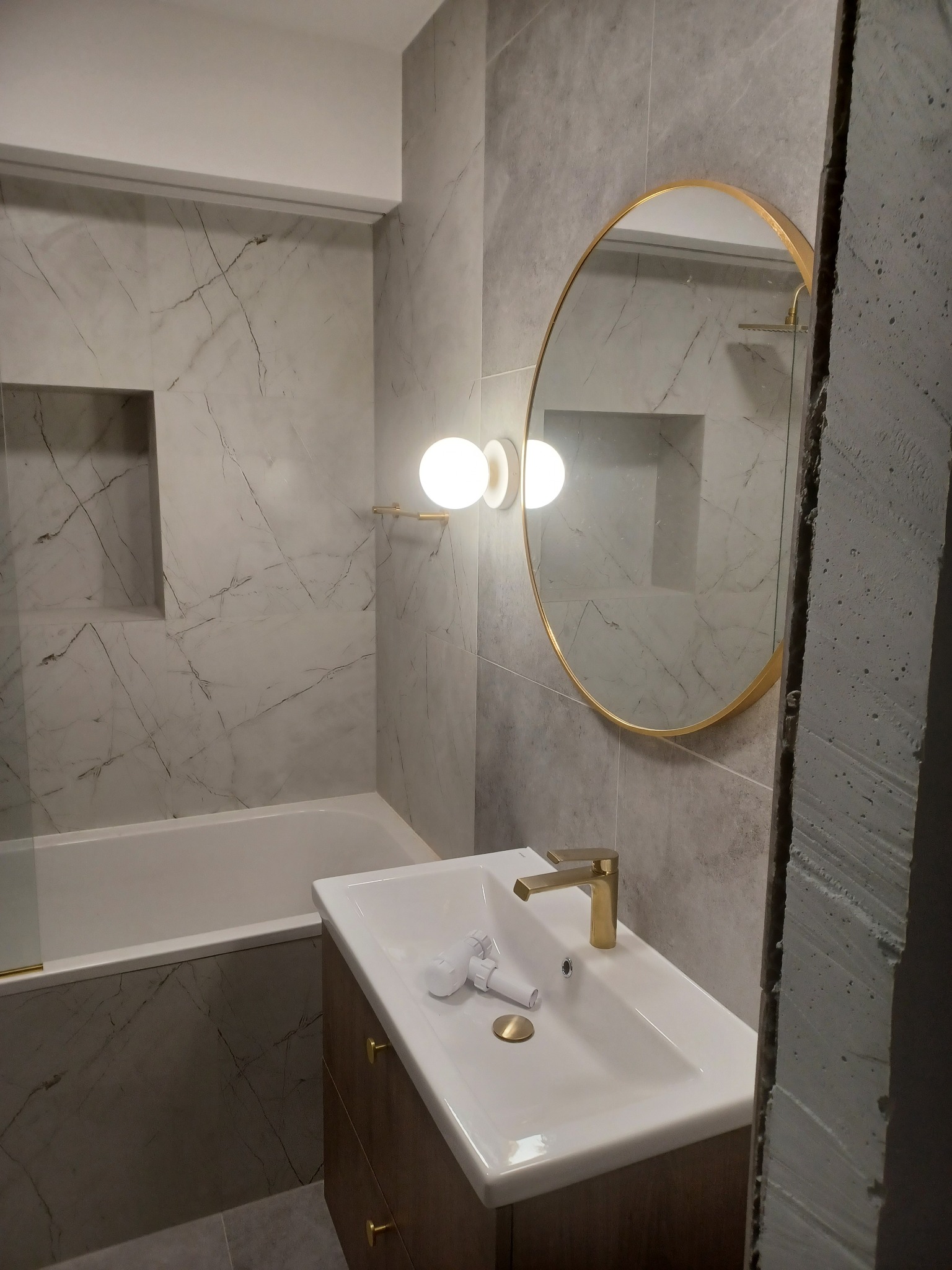 Wykończona łazienka z wanną obudowaną płytkami imitującymi marmur, umywalką nablatową na szafce z drewnopodobnymi frontami, okrągłym lustrem w złotej ramie i nowoczesnym oświetleniem.