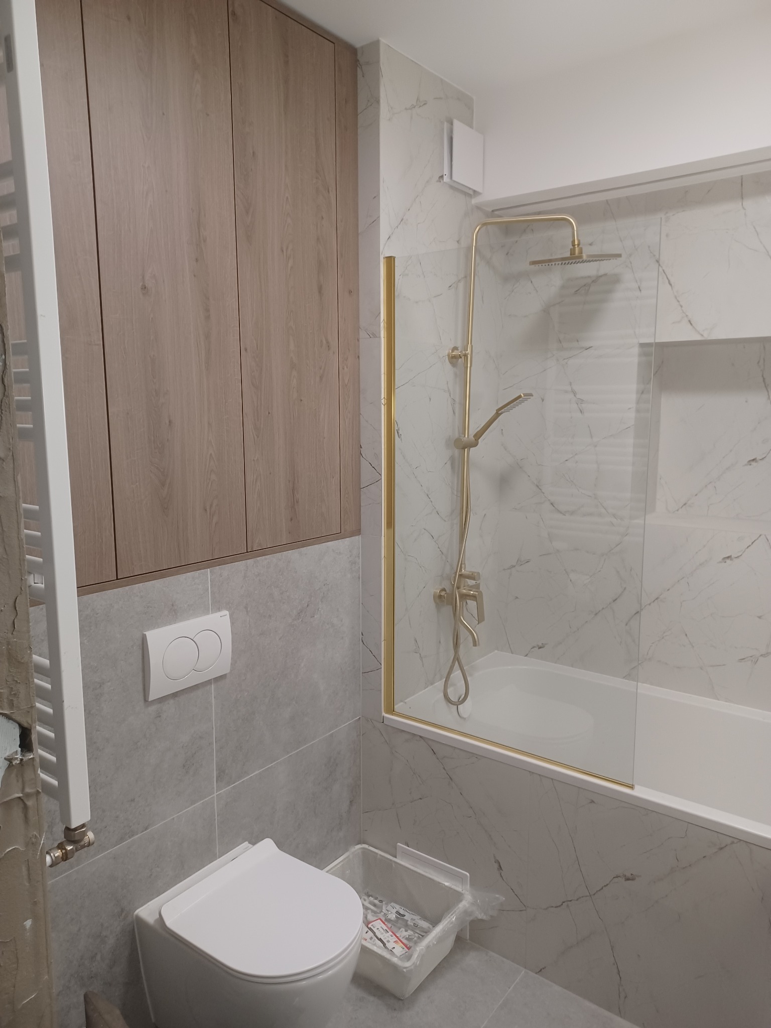 Wykończona łazienka w szaro-białej kolorystyce z marmurowym wzorem na ścianach, z zabudową szafek z drewnopodobnym fornirem i złotą armaturą prysznicową za szklaną ścianką.