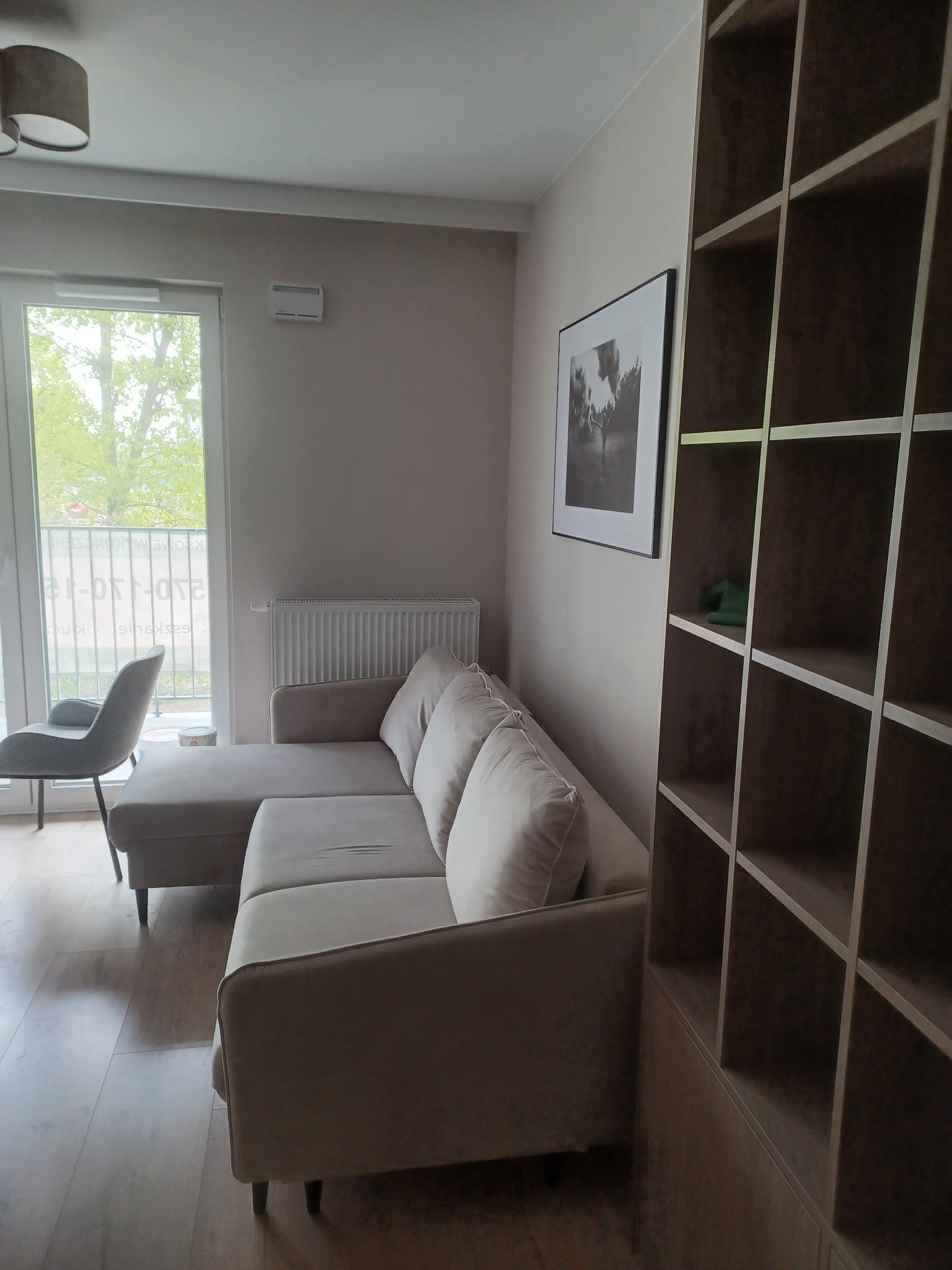 Jasny salon z narożną sofą, regałem z półkami i widokiem na balkon przez duże okno.