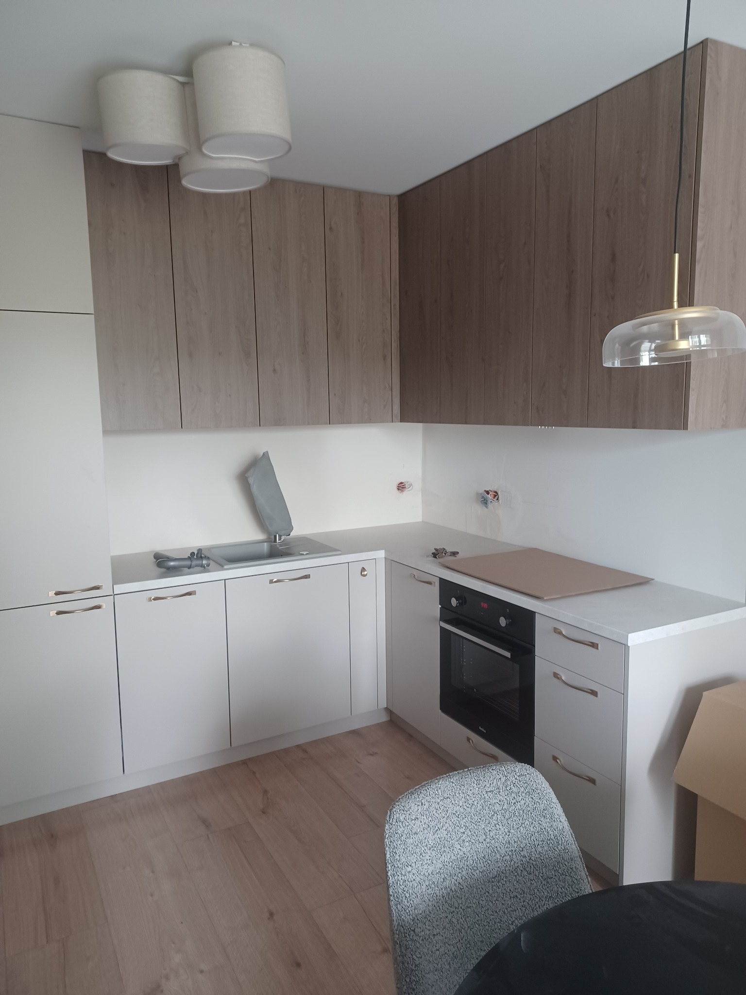 Nowoczesna kuchnia po remoncie: białe szafki z poziomymi uchwytami, drewniane szafki wiszące, płyta indukcyjna z osłoną, zlewozmywak z przykryciem, designerska lampa wisząca, parkiet na podłodze.