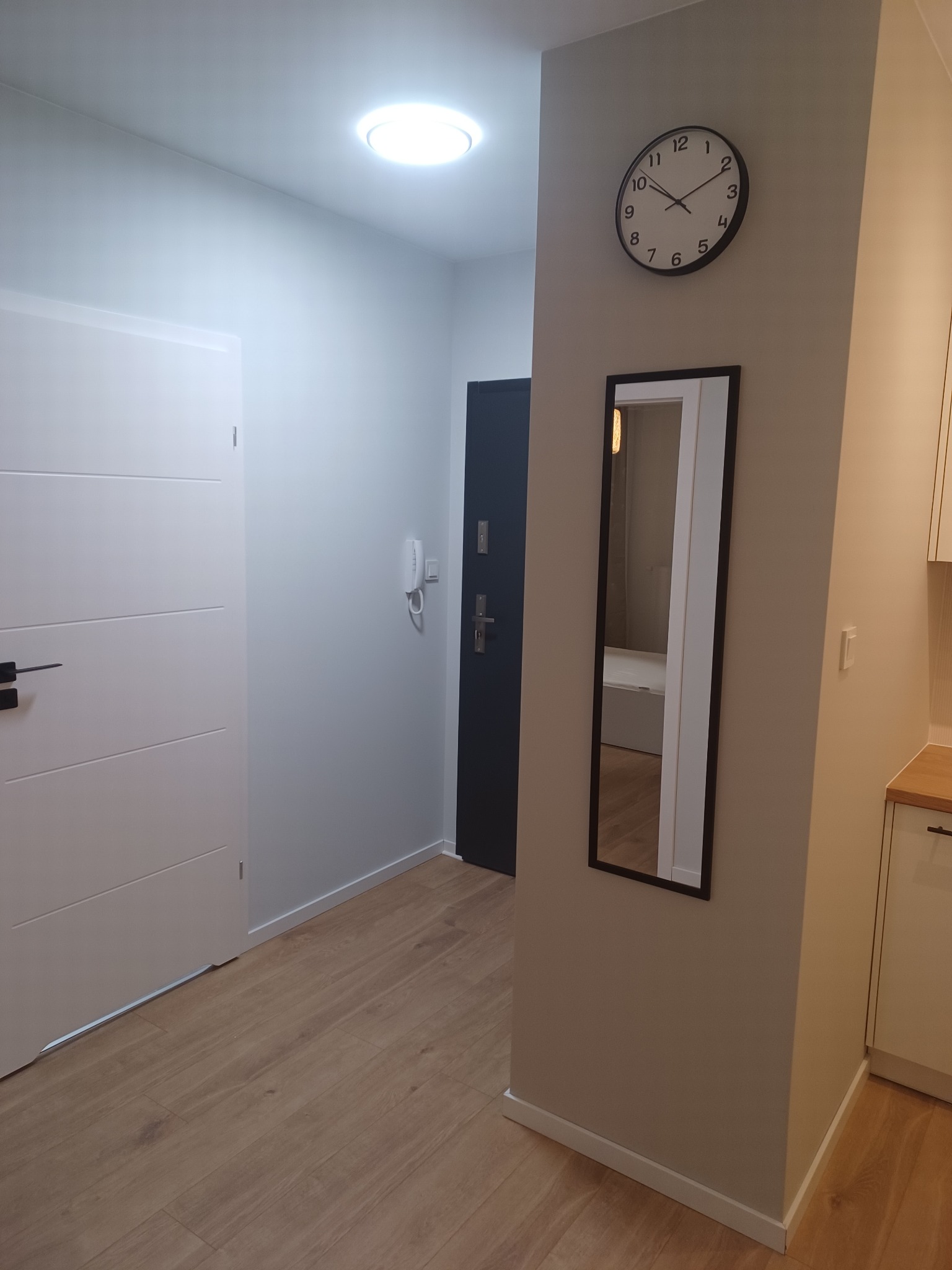 Nowoczesny przedpokój z białymi drzwiami, drewnianą podłogą, szarą ścianą z czarnym zegarem i lustrem w czarnej ramie, widok na drzwi wejściowe i fragment aneksu kuchennego.