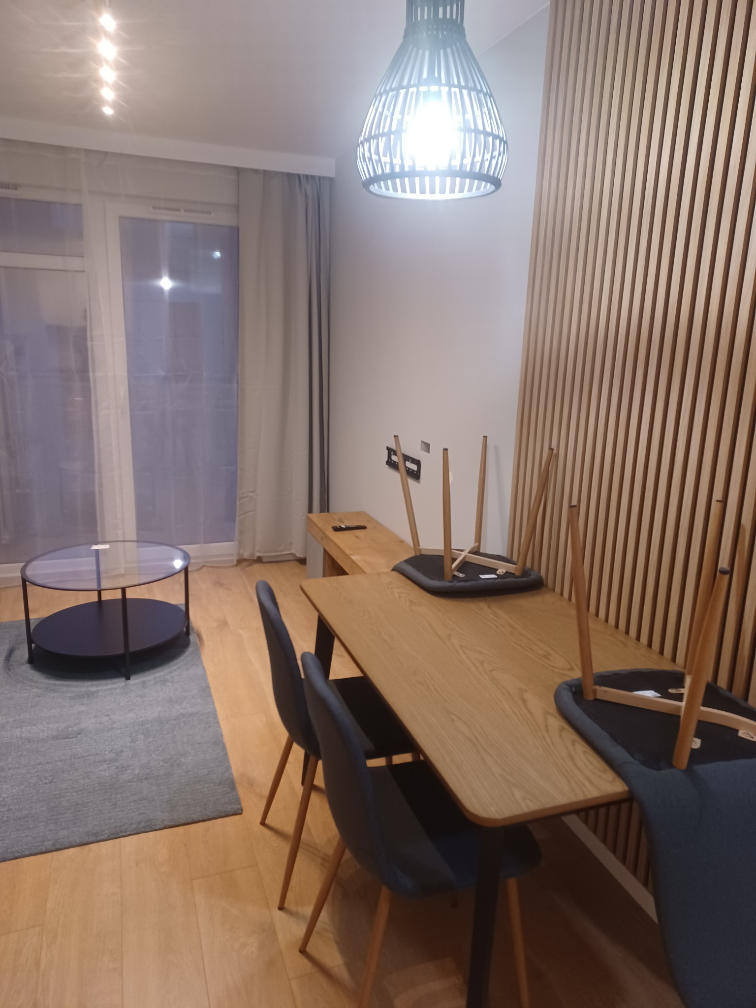 Jasne wnętrze pokoju z drewnianą ścianą, stołem z krzesłami (niektóre odwrócone do góry nogami), okrągłym stolikiem kawowym i dekoracyjną lampą wiszącą.