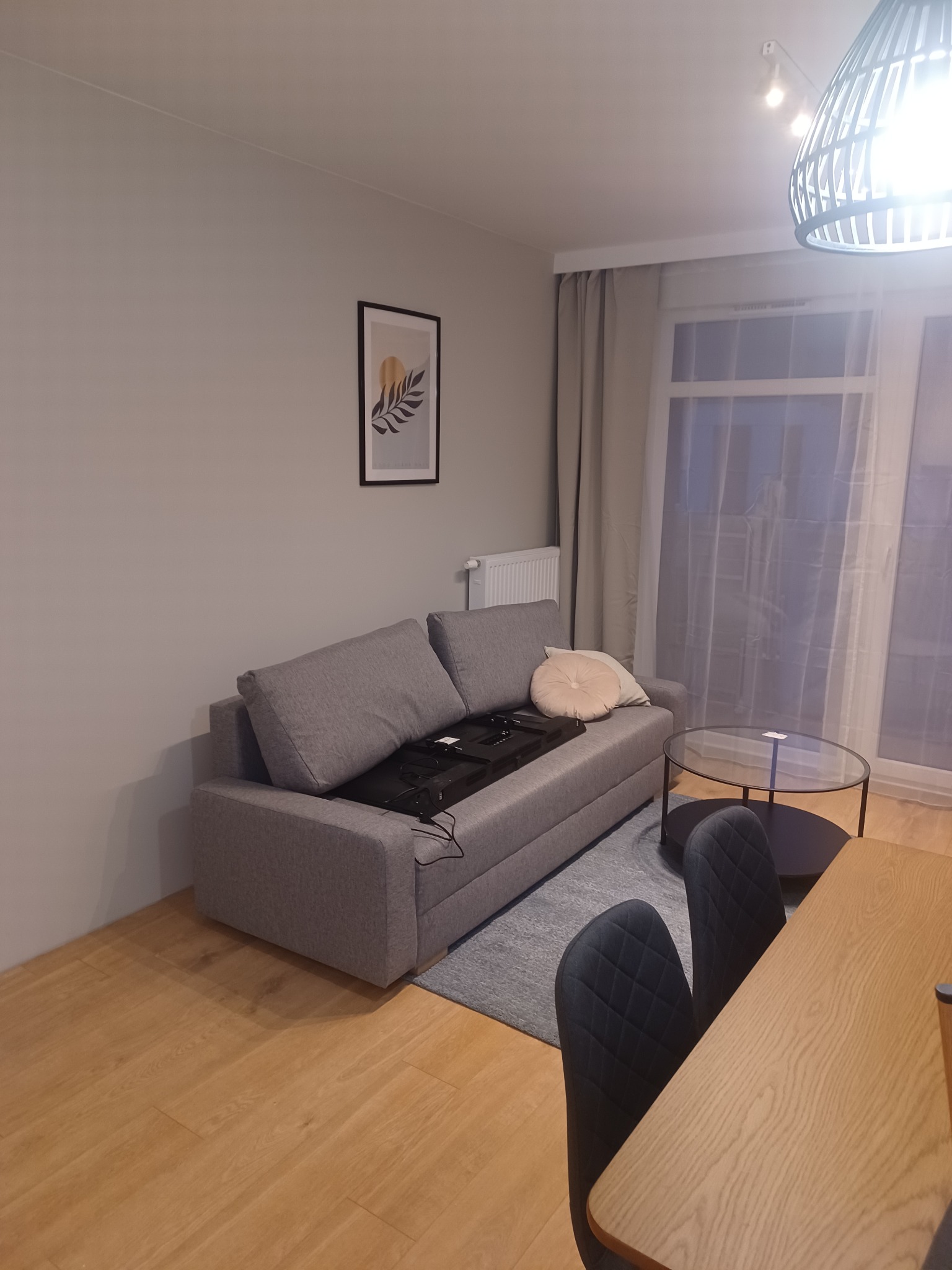 Szary rozkładany narożnik z widocznym mechanizmem rozkładania, okrągły stolik kawowy na dywanie, drewniana podłoga i minimalistyczny obrazek na ścianie w jasnym pokoju.