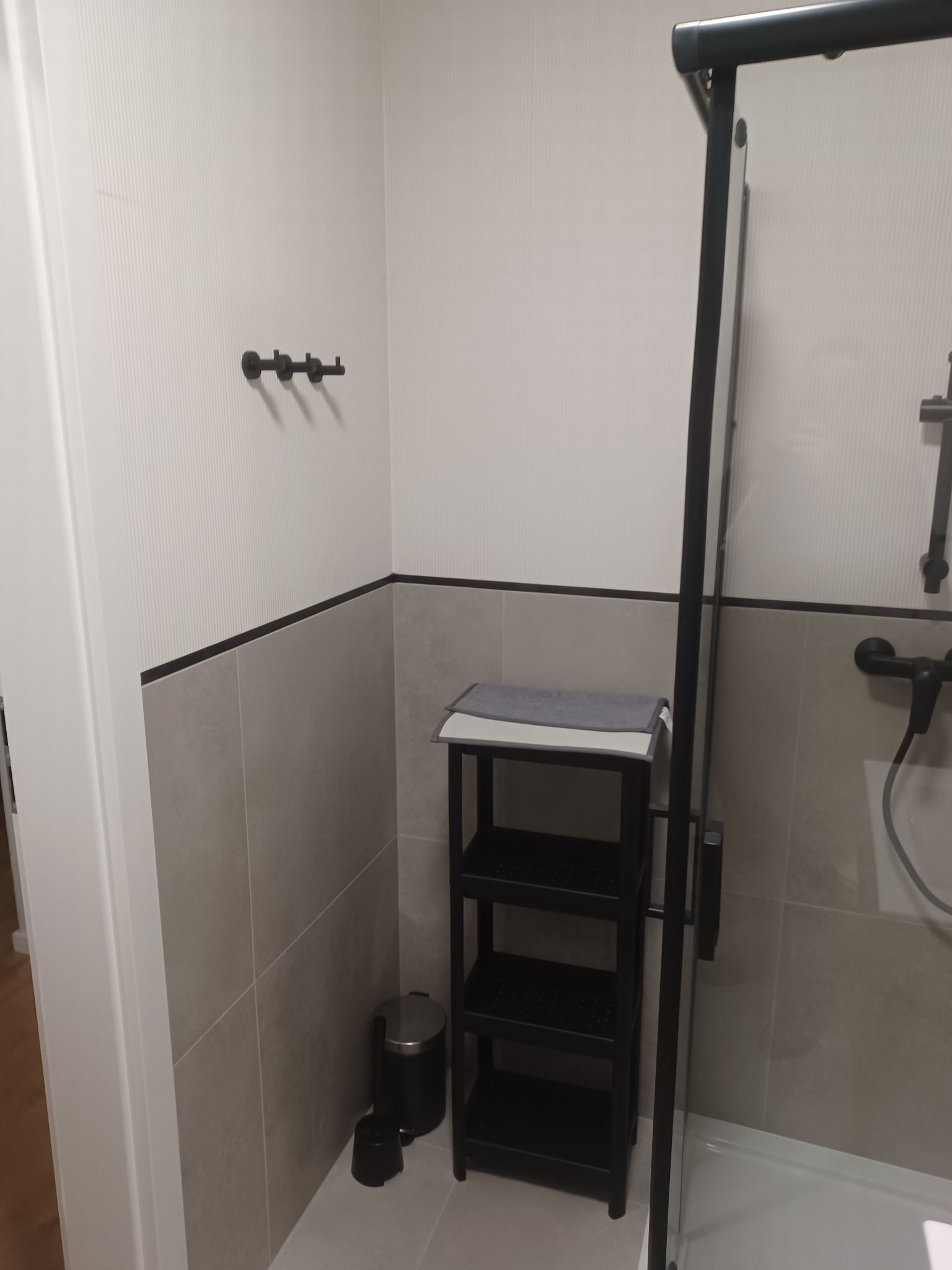 Nowoczesna łazienka z szarymi płytkami, czarnymi akcentami i minimalistycznym regałem na ręczniki obok kabiny prysznicowej.