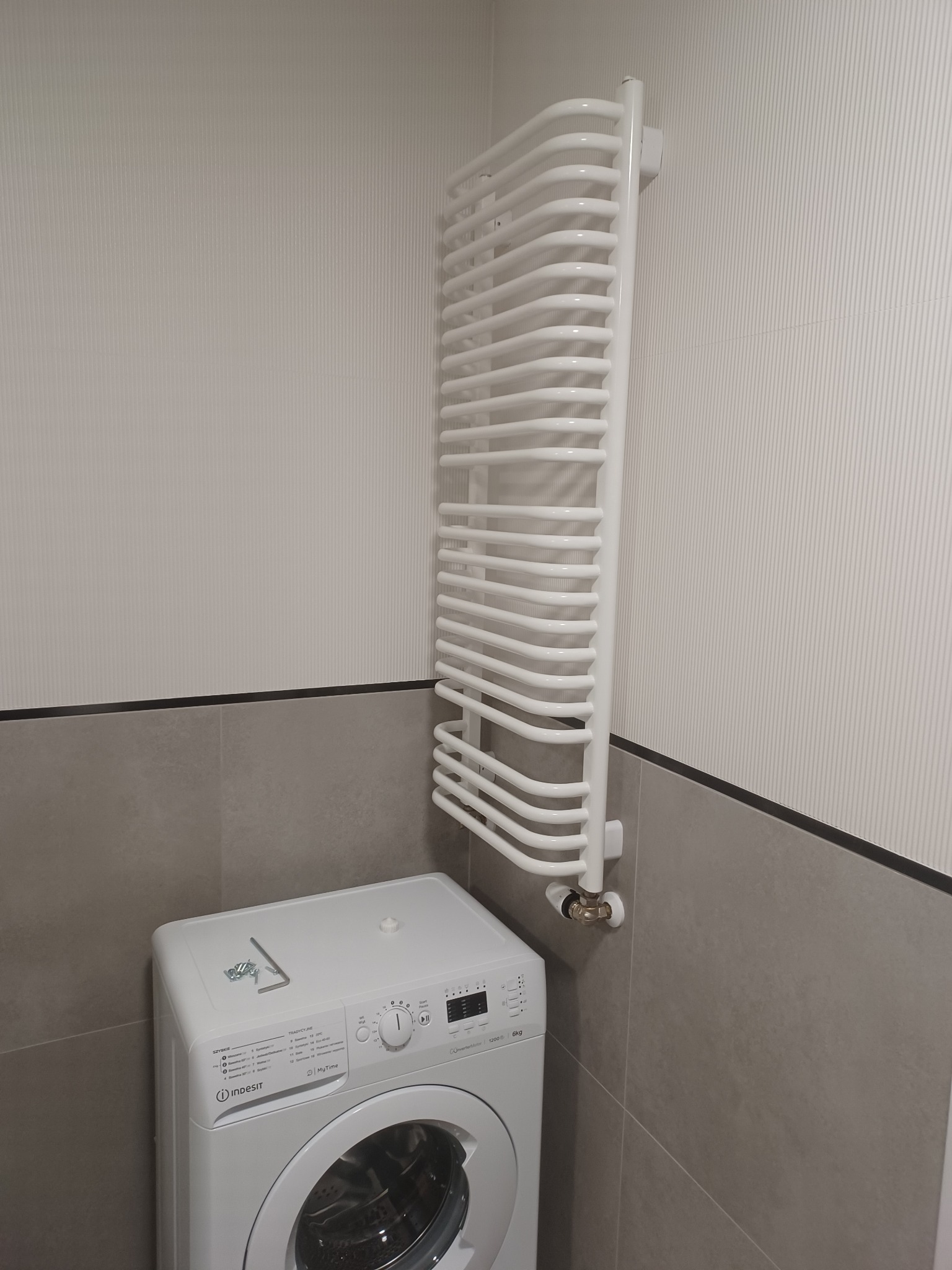 Biała pralka Indesit i grzejnik drabinkowy w łazience z szarymi płytkami i tapetą w pionowe paski.