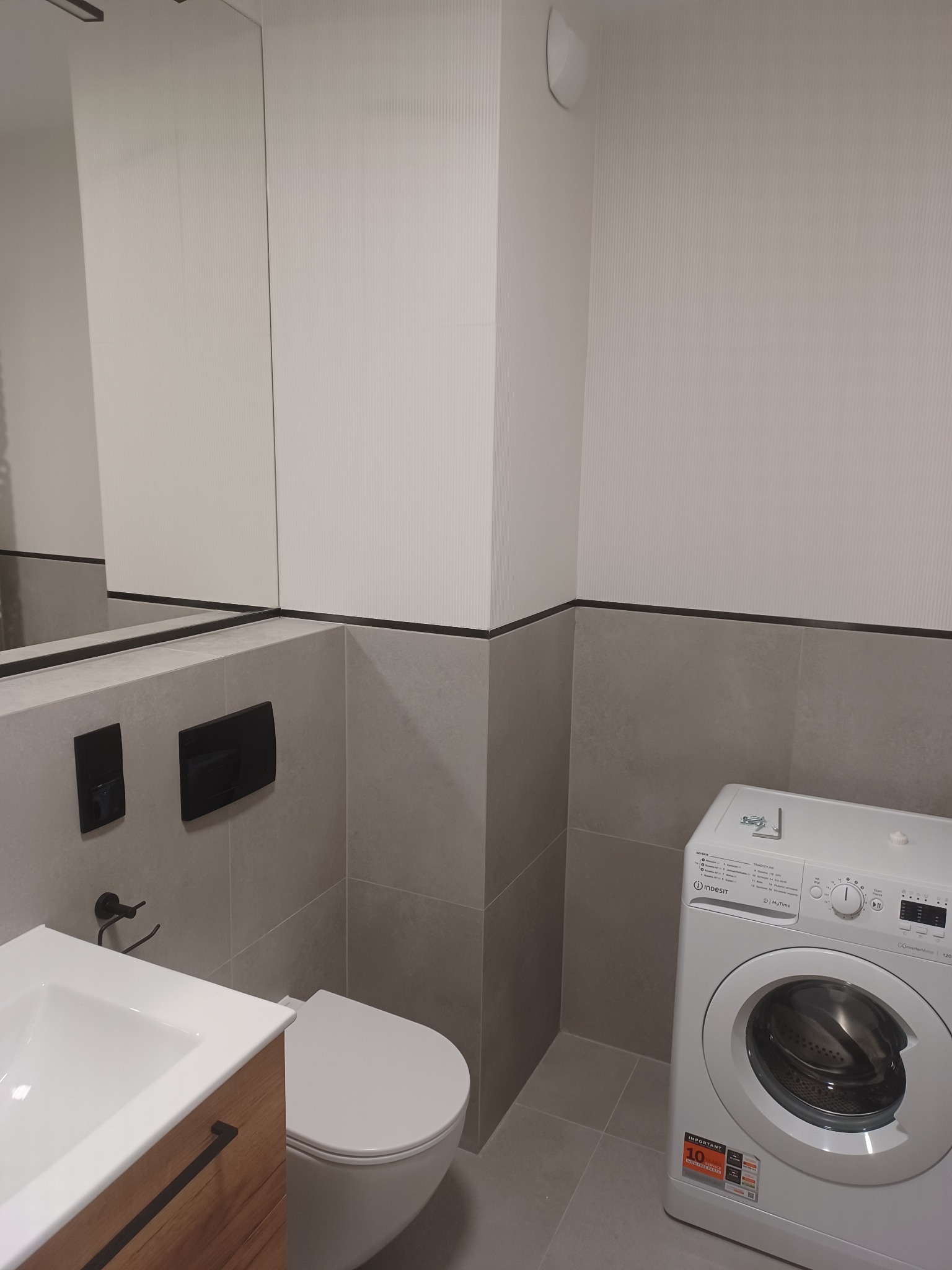 Szara łazienka po remoncie z pralką Indesit, wiszącą miską WC i umywalką z drewnianą szafką oraz czarną armaturą i przyciskami spłuczki.