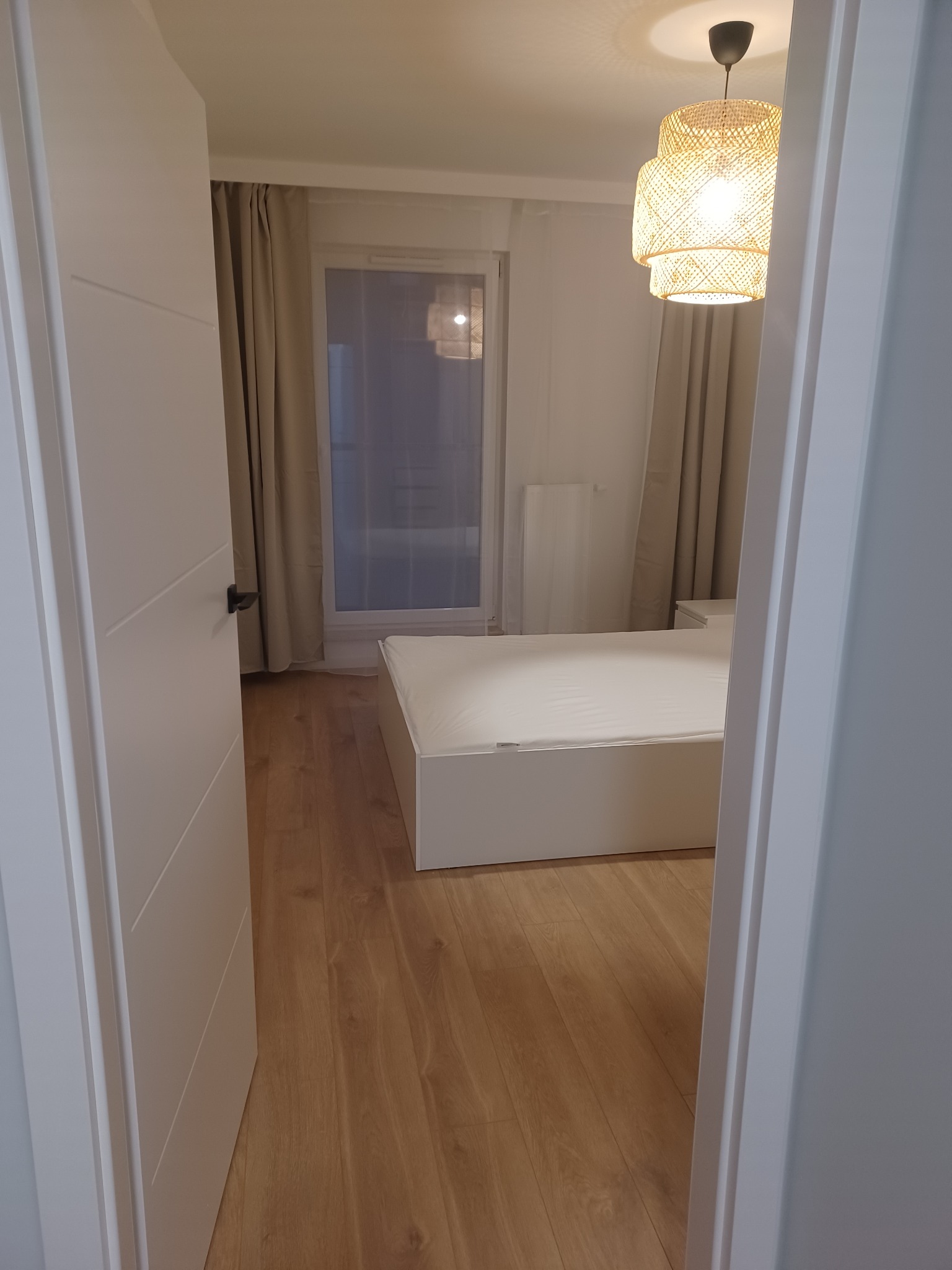 Widok na sypialnię z drewnianą podłogą, białym łóżkiem i plecioną lampą, widzianą przez otwarte białe drzwi.