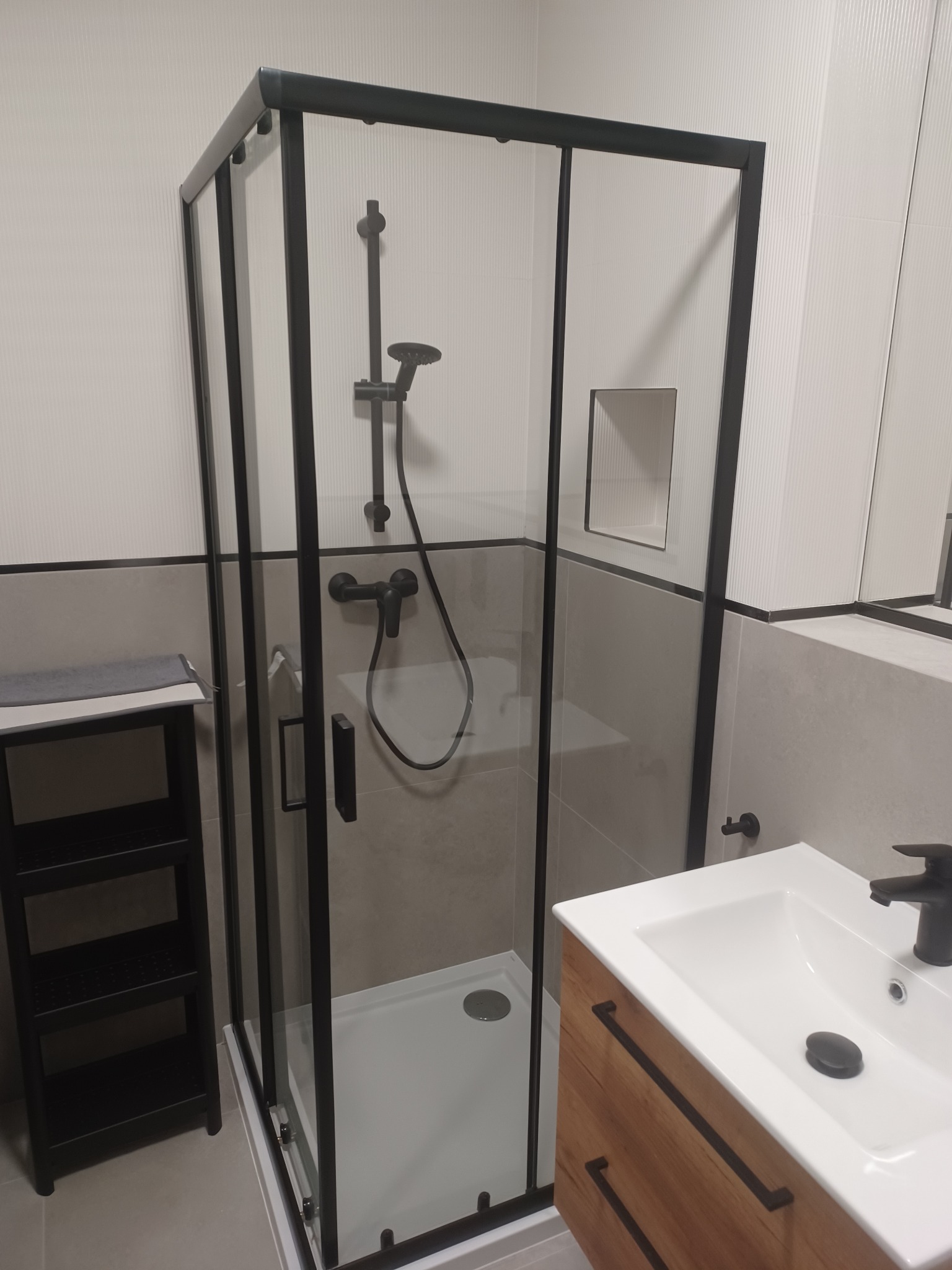 Nowoczesna kabina prysznicowa z czarnymi ramami i armaturą, umywalka z szafką z drewnopodobnym frontem, szare płytki na ścianach, minimalistyczny design łazienki po remoncie.