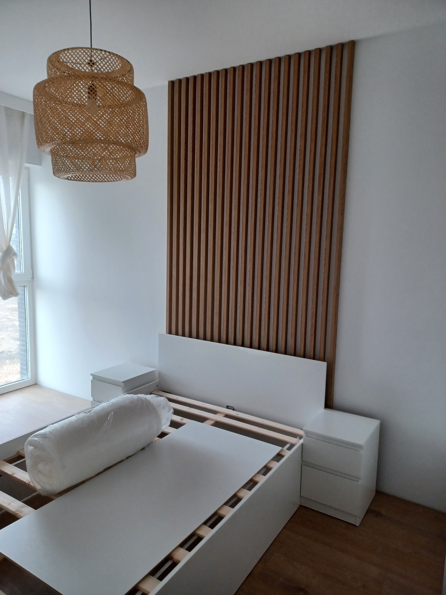 Montaż drewnianego panelu dekoracyjnego za łóżkiem w sypialni, widoczna lampa z wikliny, stelaż łóżka i szafki nocne w minimalistycznym stylu.