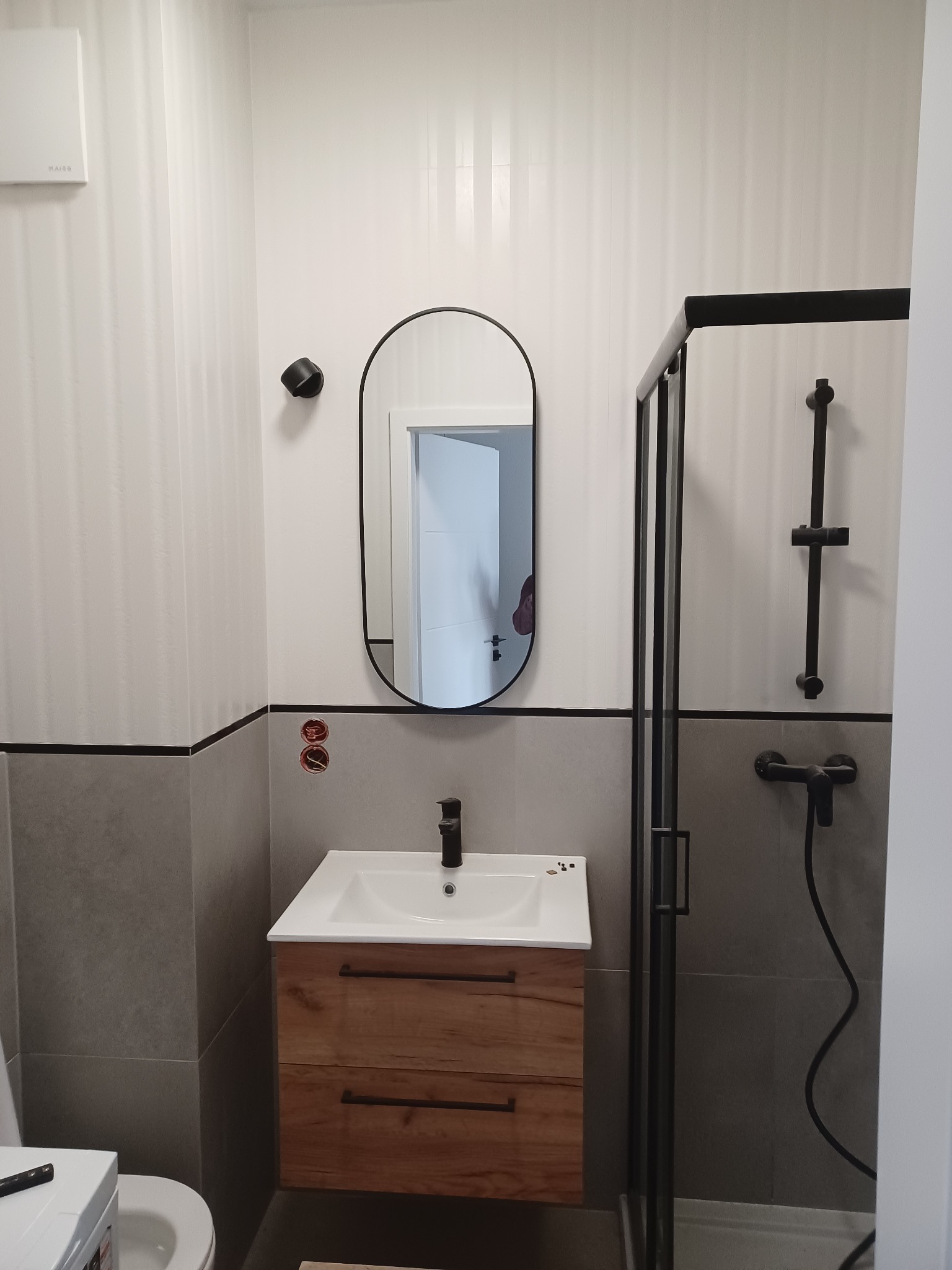 Nowoczesna łazienka z umywalką na drewnianej szafce, owalnym lustrem w czarnej ramie i kabiną prysznicową z czarnymi profilami, wykończona płytkami w odcieniach szarości i bieli, z widocznymi...