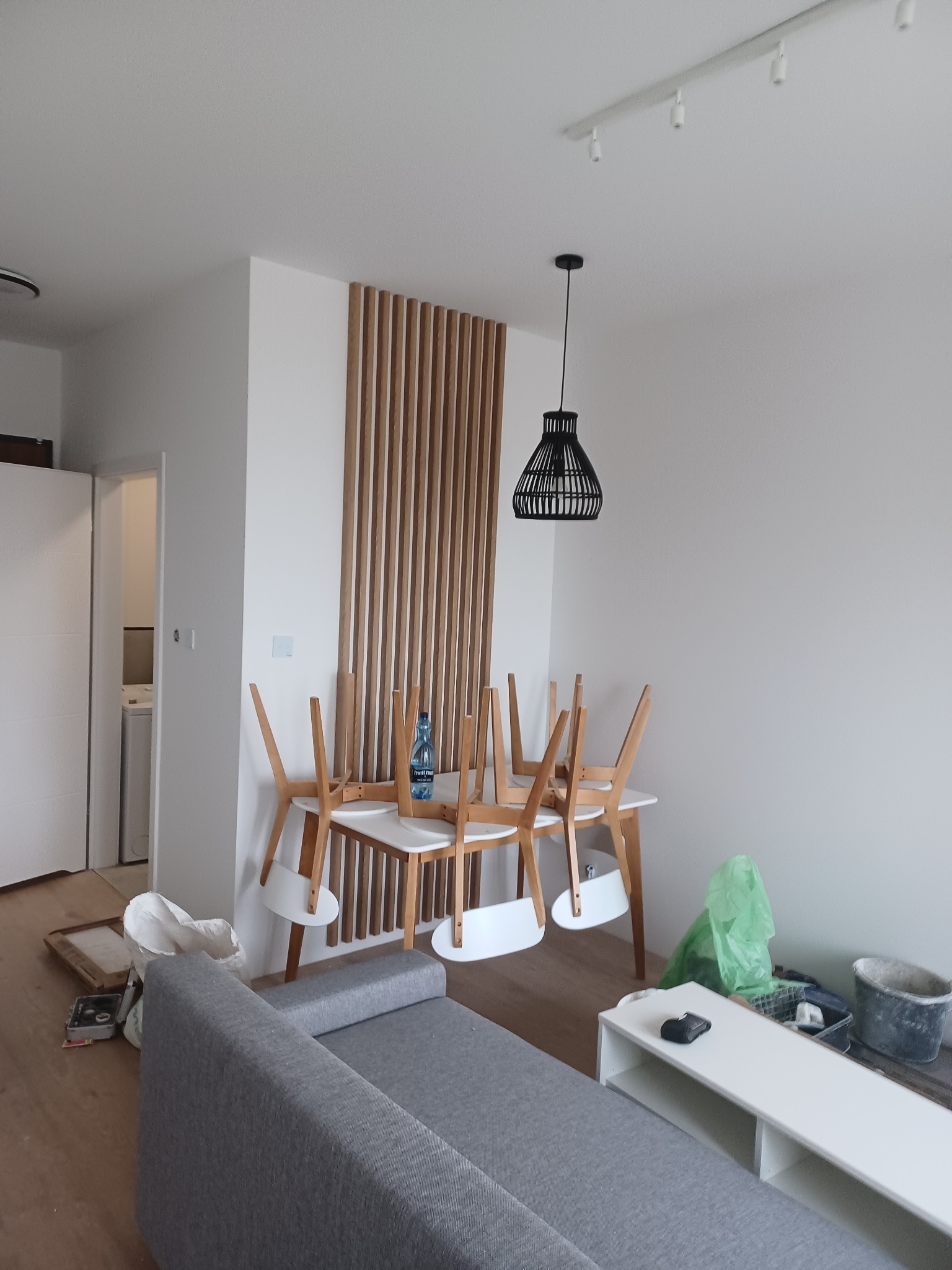Wnętrze mieszkania w trakcie remontu: szary narożnik, stół z odwróconymi krzesłami, butelka wody, biała komoda, lampa wisząca, akcenty drewniane, widoczne narzędzia i materiały remontowe.