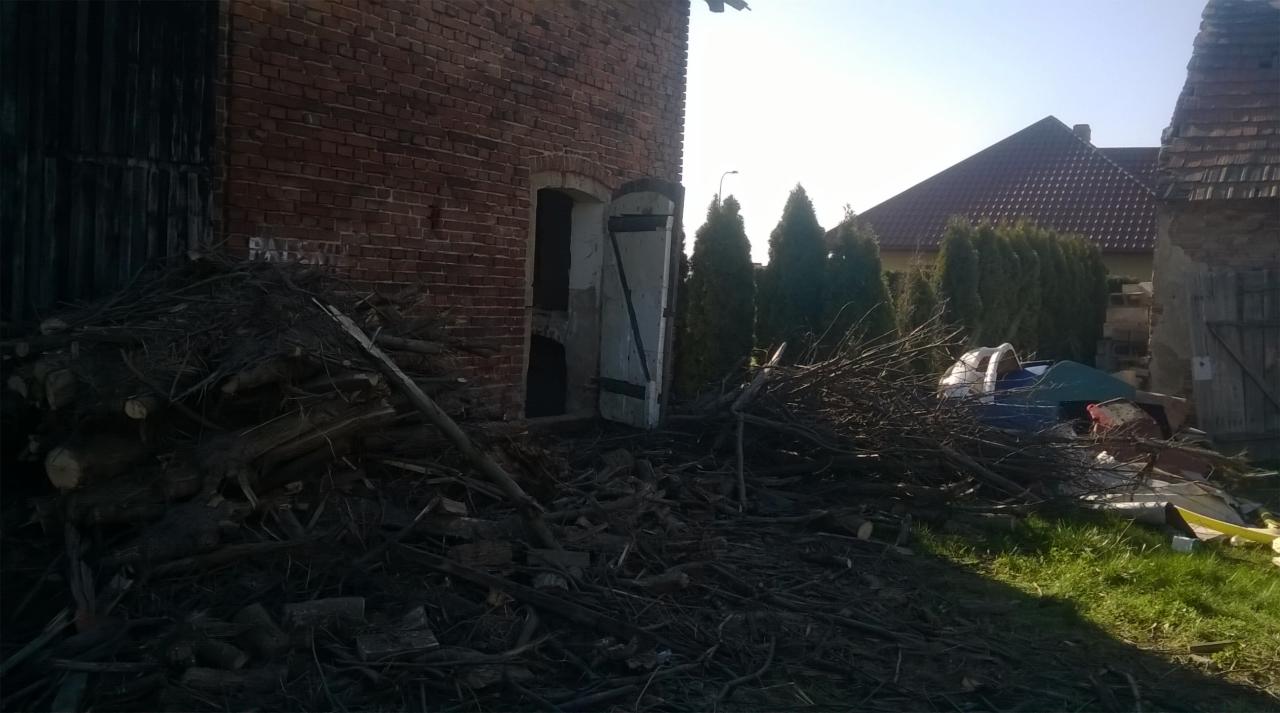 Stary, ceglany budynek gospodarczy z otwartymi, białymi drzwiami i stertą gałęzi oraz śmieci na pierwszym planie, widoczne fragmenty innych budynków w tle.