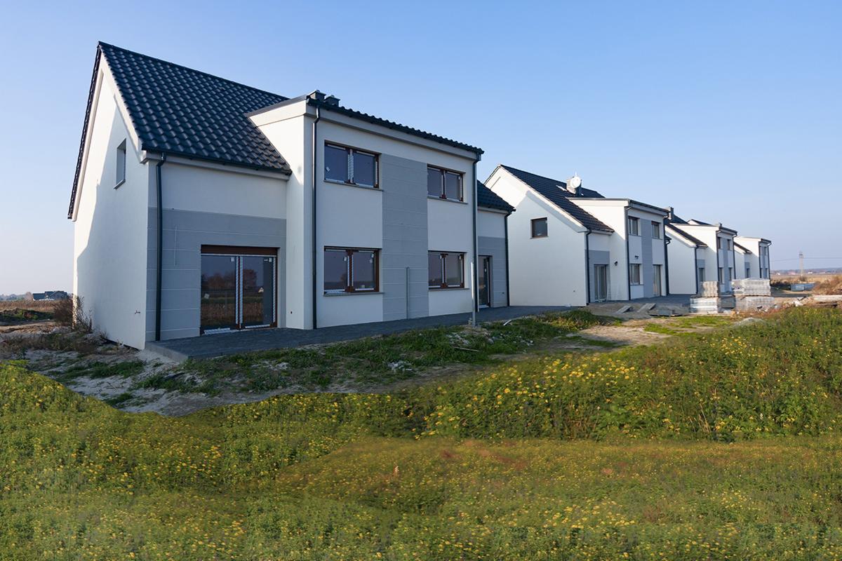 Szereg nowo wybudowanych, dwukondygnacyjnych domów z ciemną dachówką i jasną elewacją z szarymi akcentami, widocznych z perspektywy trawnika z żółtymi kwiatami.