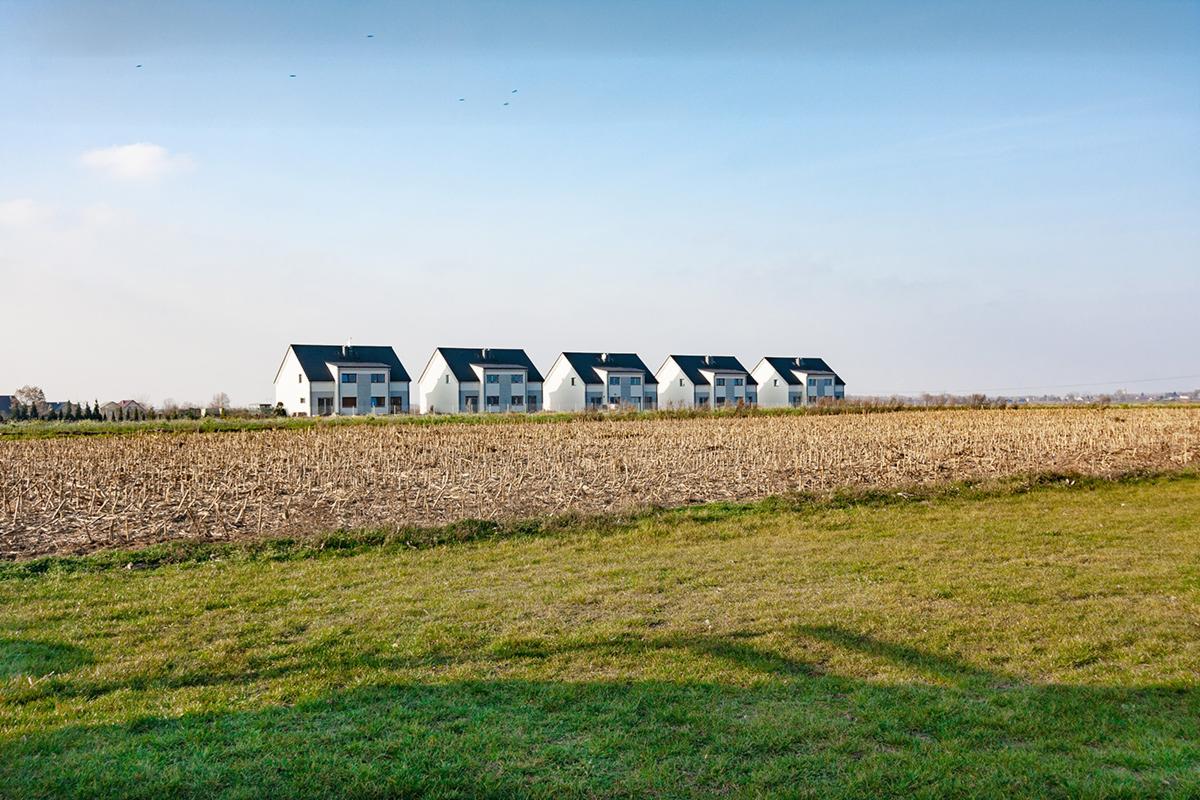 Rząd sześciu identycznych, nowoczesnych domów z ciemnymi dachami, stojących na skraju pola uprawnego, widok z daleka w słoneczny dzień.