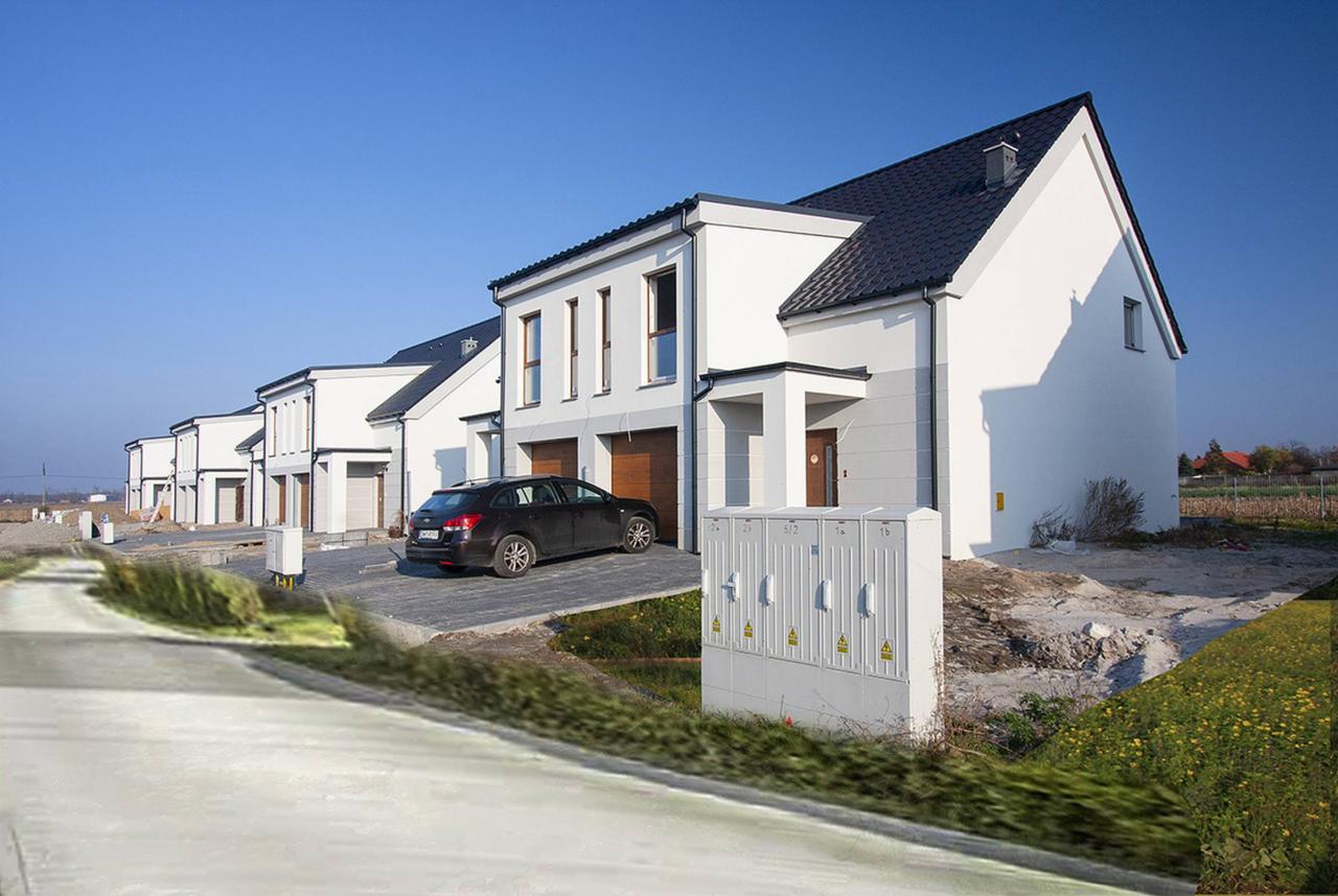 Szereg identycznych, nowoczesnych domów jednorodzinnych z garażami, zaparkowany samochód osobowy na podjeździe z kostki brukowej, skrzynki elektryczne na pierwszym planie.