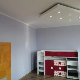 Świeżo pomalowane na liliowo ściany pokoju z piecem kaflowym w kolorze miodowym, białym sufitem z wbudowanymi punktowymi lampami i meblami dziecięcymi z elementami w kolorze czerwonym.