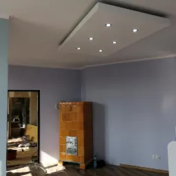 Pokój po remoncie z pomalowanymi ścianami na fioletowo-szaro, z widocznym piecem kaflowym i podwieszanym sufitem z oświetleniem LED. W tle widoczne pomieszczenie w trakcie remontu.