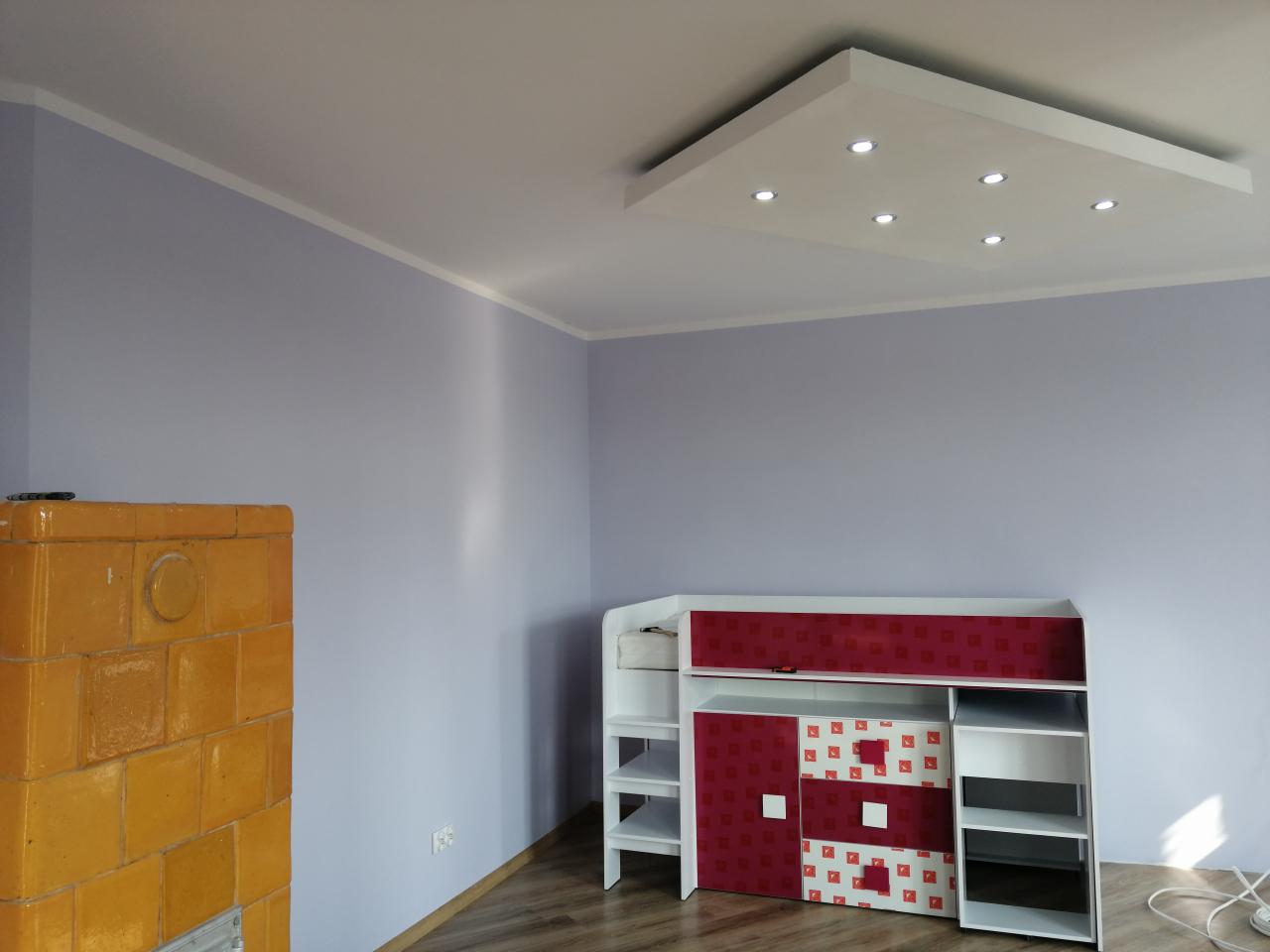 Świeżo pomalowane na liliowo ściany pokoju z piecem kaflowym w kolorze miodowym, białym sufitem z wbudowanymi punktowymi lampami i meblami dziecięcymi z elementami w kolorze czerwonym.