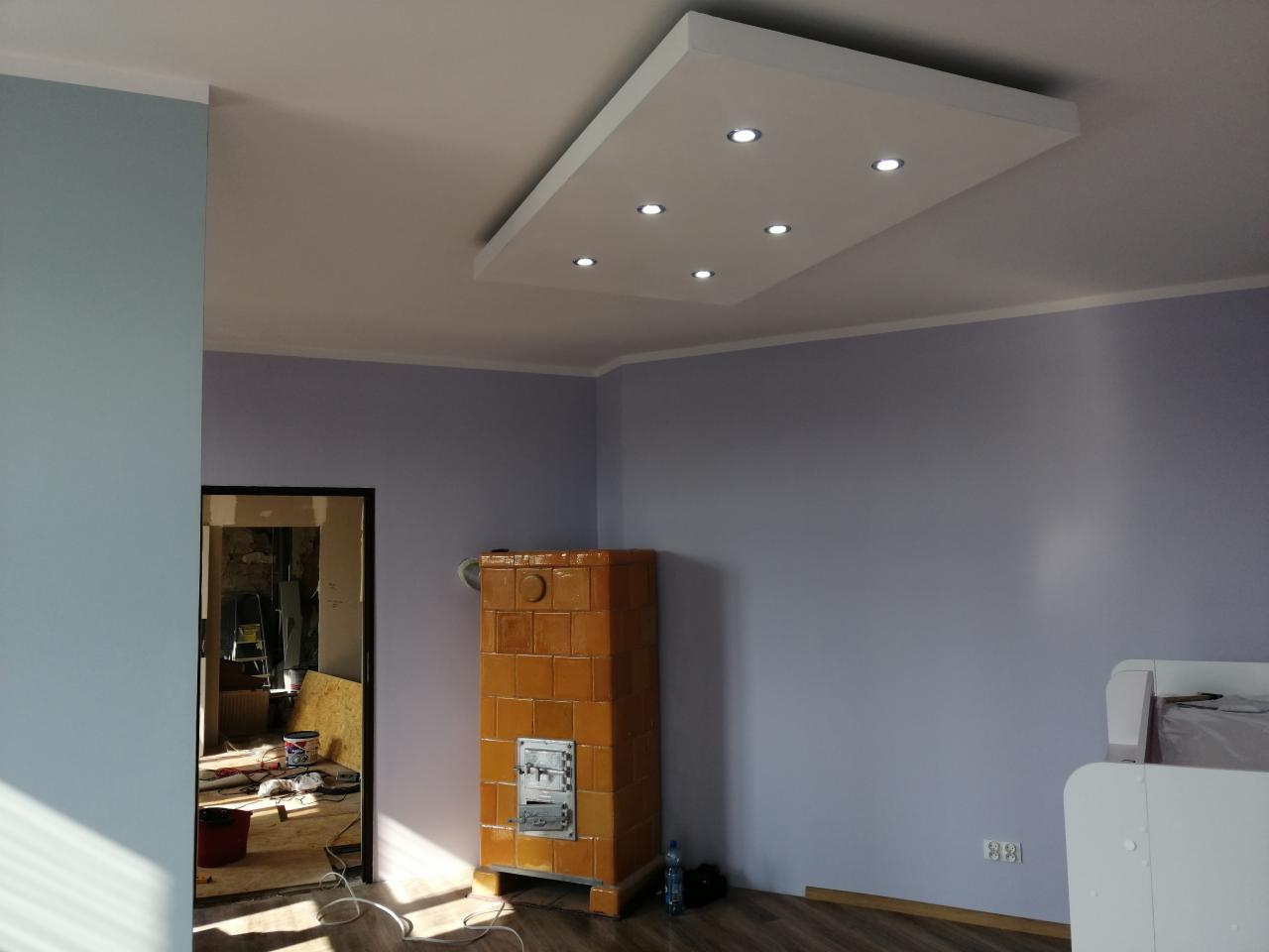 Pokój po remoncie z pomalowanymi ścianami na fioletowo-szaro, z widocznym piecem kaflowym i podwieszanym sufitem z oświetleniem LED. W tle widoczne pomieszczenie w trakcie remontu.