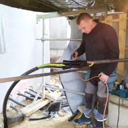 Elektryk łączy kable elektryczne w surowym wnętrzu budynku w trakcie budowy, widoczne narzędzia i elementy konstrukcyjne.