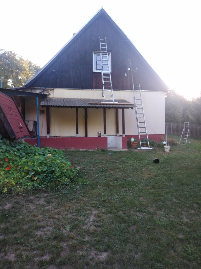 Dom jednorodzinny z drewnianą elewacją szczytową i dwiema rozstawionymi drabinami aluminiowymi, przygotowany do prac na wysokości.