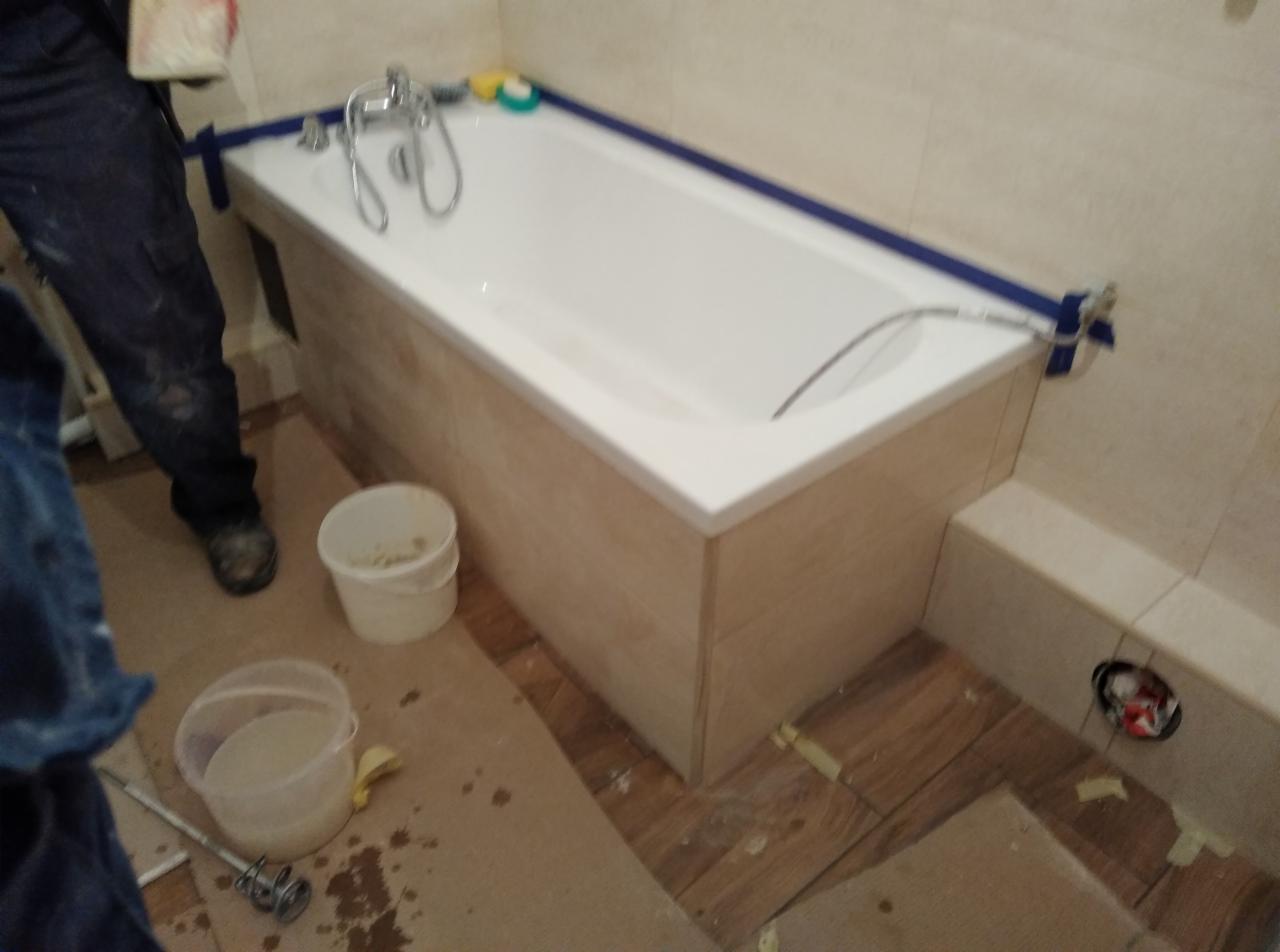Obudowa wanny wykończona płytkami imitującymi drewno, zabezpieczona niebieską taśmą malarską, widoczne narzędzia i materiały budowlane, etap prac wykończeniowych w łazience.