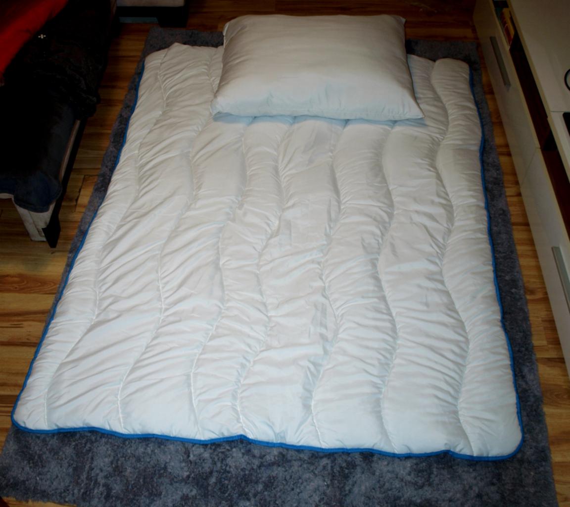 Biała kołdra z niebieską lamówką i poduszka leżą na szarym dywanie shaggy na drewnianej podłodze, widoczny fragment mebli.