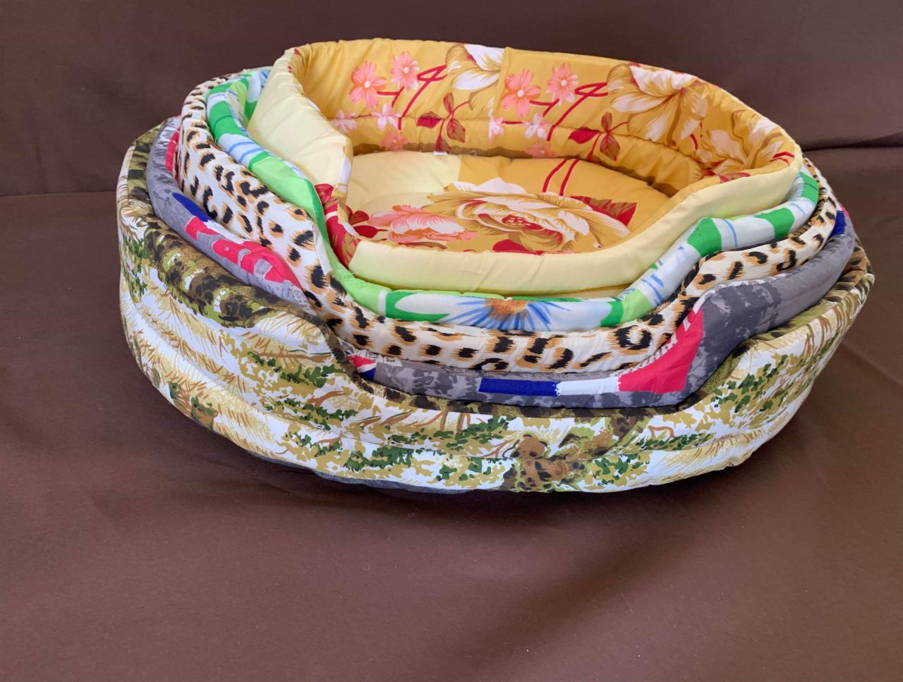 Stos pięciu legowisk dla zwierząt z różnorodnych, wzorzystych tkanin, od kwiatowych po zwierzęce printy, ułożonych jedno w drugim na brązowym tle.