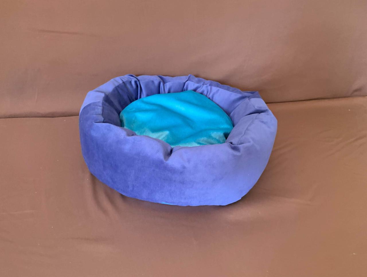 Okrągłe, fioletowe legowisko dla zwierząt z turkusową poduszką w środku, umieszczone na brązowym tle.