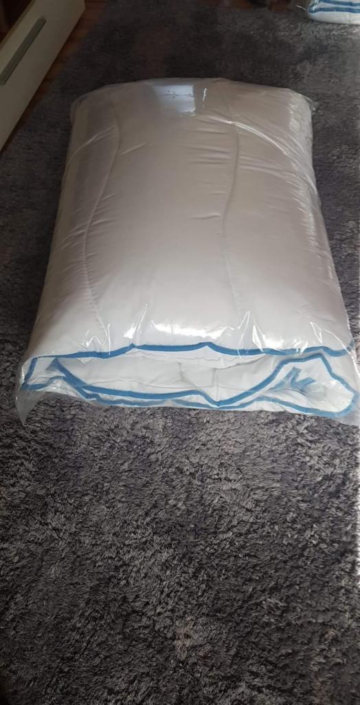 Biała kołdra w przezroczystym opakowaniu foliowym, z niebieskim zygzakowatym obszyciem, leżąca na szarym dywanie.