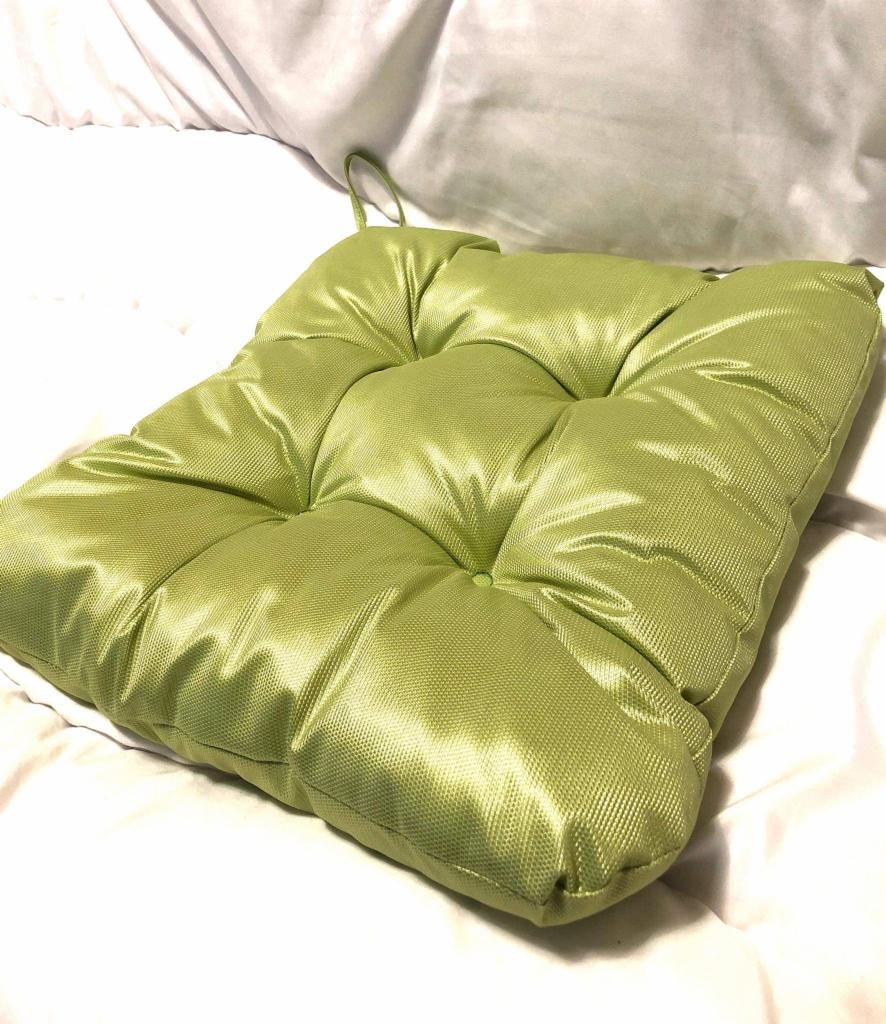 Pikowana, jasnozielona poduszka na krzesło z połyskującej tkaniny, z uchwytem do zawieszenia, leżąca na białej pościeli.