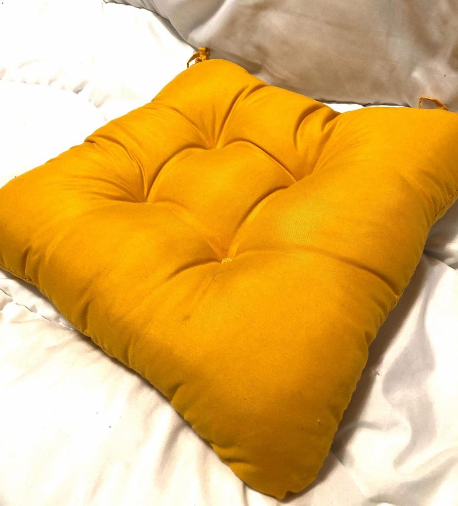 Żółta, kwadratowa poduszka na krzesło z guzikami, leżąca na białej pościeli.