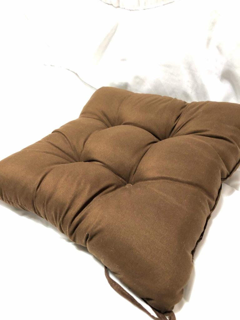 Kwadratowa, brązowa poduszka na krzesło z wiązaniami, leżąca na białym materiale.