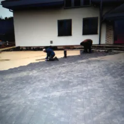 Dwaj pracownicy układają ciemnoszarą kostkę brukową na podsypce piaskowej przed domem z białymi ścianami i ciemnymi oknami; jeden z pracowników klęczy, wyrównując powierzchnię.