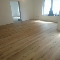 Jasne panele podłogowe imitujące drewno w pustym pokoju z białymi ścianami, widoczne przygotowania do wykończenia wnętrza w sąsiednim pomieszczeniu.