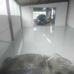 Świeżo wylana, jasnoszara posadzka w warsztacie samochodowym, widoczne narzędzia i samochód w tle.