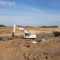 Biała koparka Liebherr pracuje w wykopie ziemnym, widoczni pracownicy w kaskach, krajobraz z nasypem w tle, błękitne niebo.