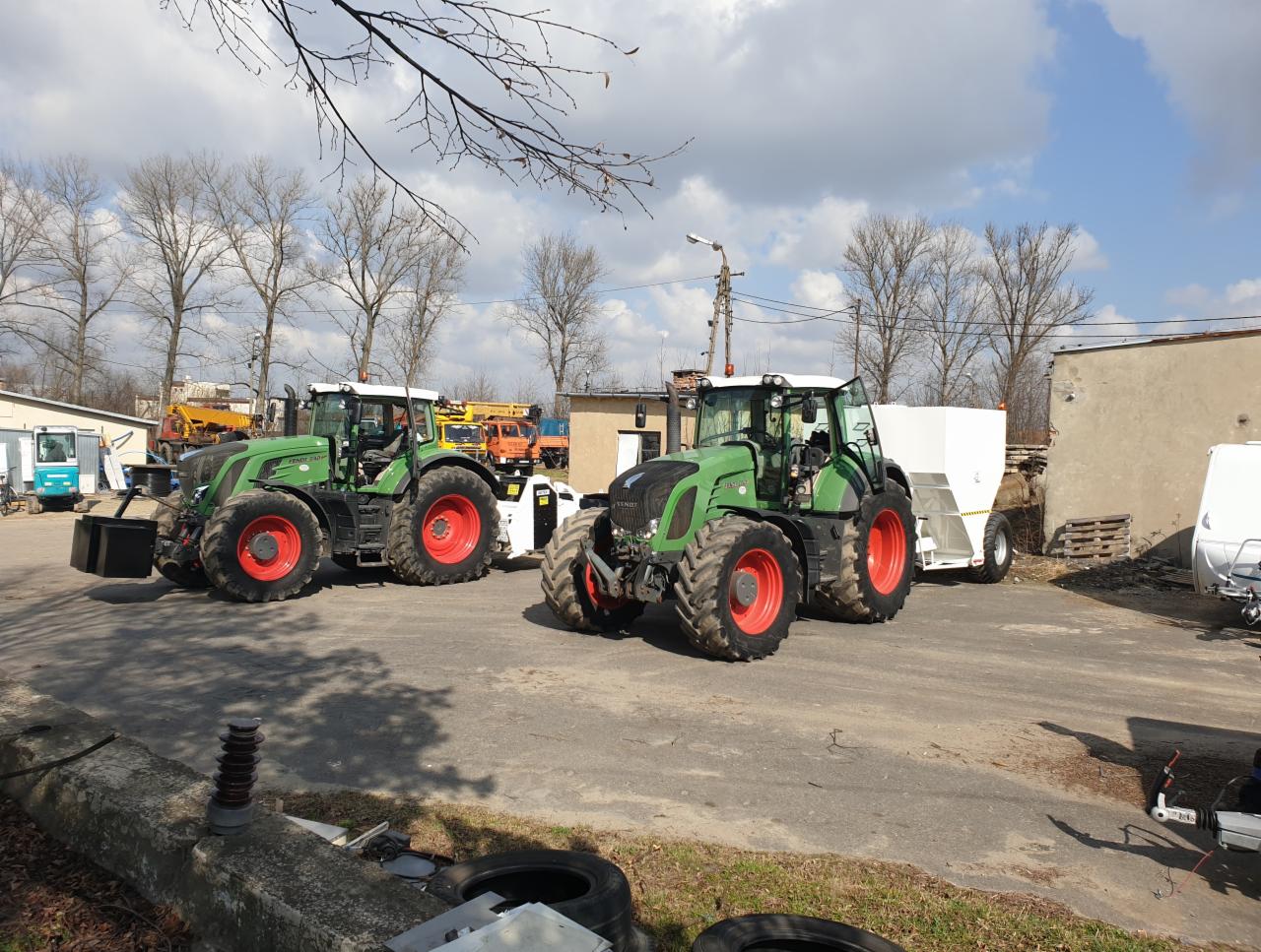 Dwa zielone traktory Fendt z czerwonymi felgami, jeden z przednim obciążnikiem, drugi z białą przyczepą, stojące na placu w słoneczny dzień.