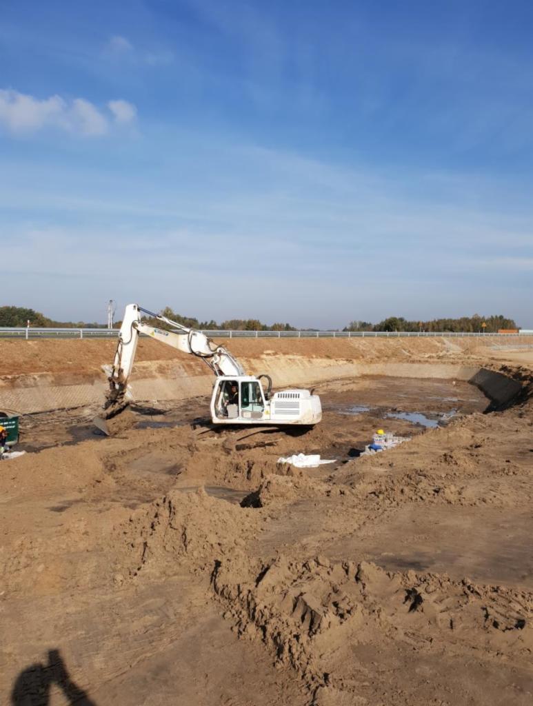 Biała koparka Liebherr pracuje w wykopie ziemnym, widoczni pracownicy w kaskach, krajobraz z nasypem w tle, błękitne niebo.