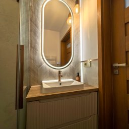 Nowoczesna łazienka z umywalką nablatową, podświetlanym lustrem w kształcie owalu i drewnianymi akcentami. Ściana wyłożona płytkami imitującymi drewno w jodełkę.