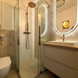 Nowoczesna łazienka z kabiną prysznicową typu walk-in, umywalką na szafce z frezowanymi frontami i podświetlanym lustrem. Uwagę zwracają złote detale armatury.