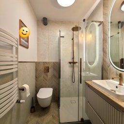 Nowoczesna łazienka z kabiną prysznicową typu walk-in, umywalką na szafce z drewnianym blatem i grzejnikiem drabinkowym. Uwagę zwracają płytki ułożone w jodełkę na podłodze.