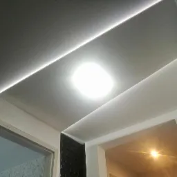 Sufit podwieszany z oświetleniem LED wzdłuż krawędzi, z fragmentem ściany pokrytej czarną, tr&oacute;jwymiarową okładziną i widocznym fragmentem ościeżnicy drzwiowej.