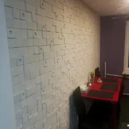 Ściana z dekoracyjnych, trójwymiarowych paneli gipsowych w jadalni z czerwonym stołem i czarnymi krzesłami.