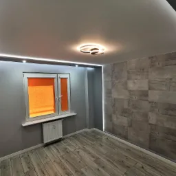 Wykończone wnętrze pokoju: szare ściany, panelowa ściana z imitacją kamienia z podświetleniem LED, drewnopodobna podłoga, okno z pomarańczową roletą i nowoczesna lampa sufitowa