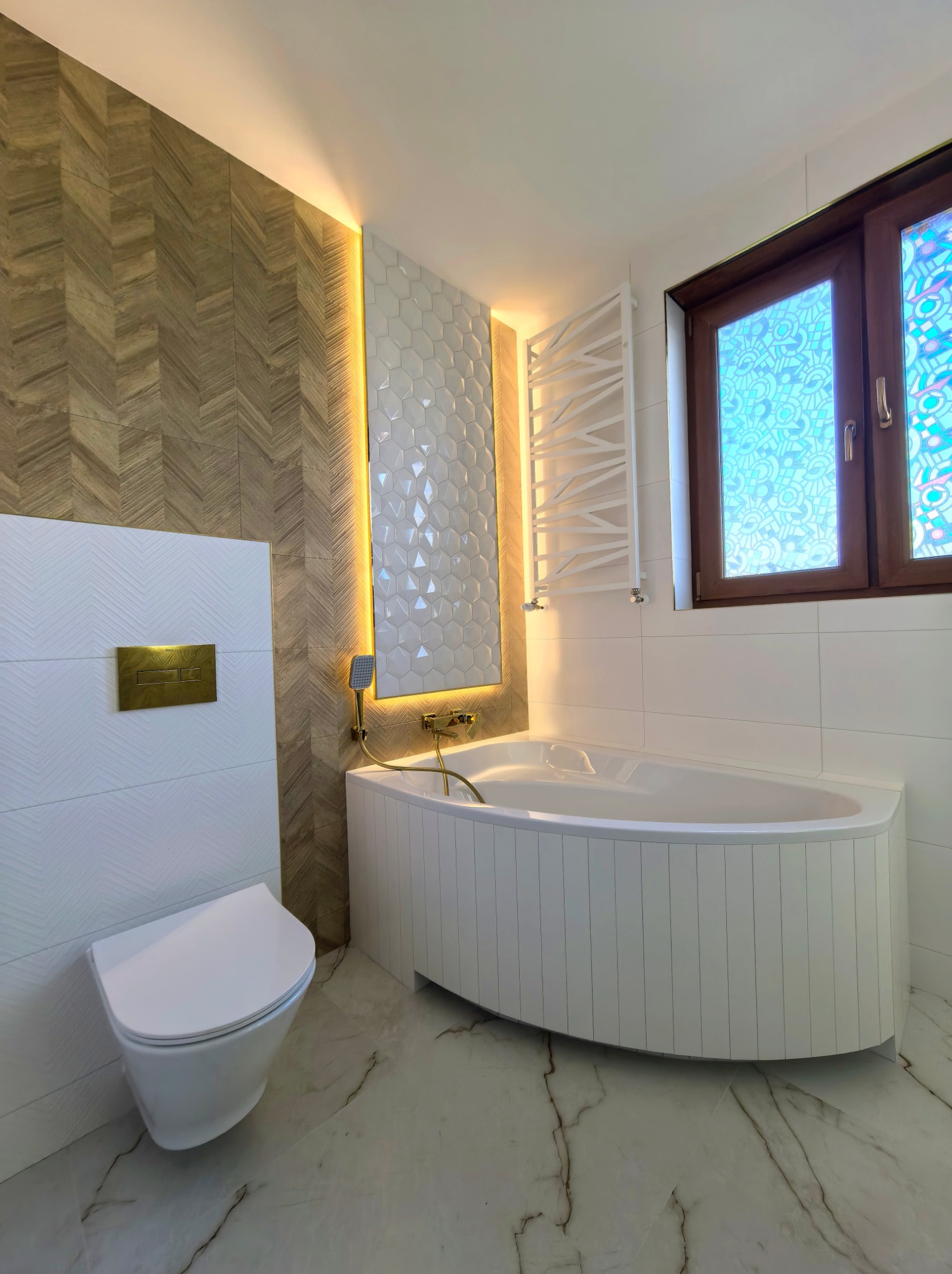 Nowoczesna łazienka z wanną, toaletą podwieszaną i drewnopodobnymi płytkami ułożonymi w jodełkę, oświetlona ciepłym światłem LED.