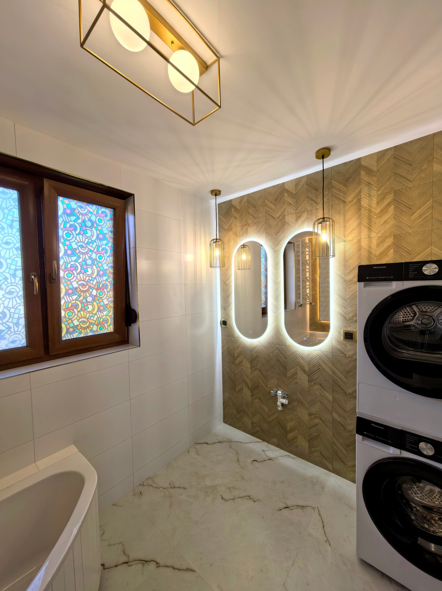 Nowoczesna łazienka z drewnianą mozaiką na ścianie, podświetlanymi lustrami i pralką z suszarką. Okno z kolorową folią. Marmurowa podłoga i złote akcenty.
