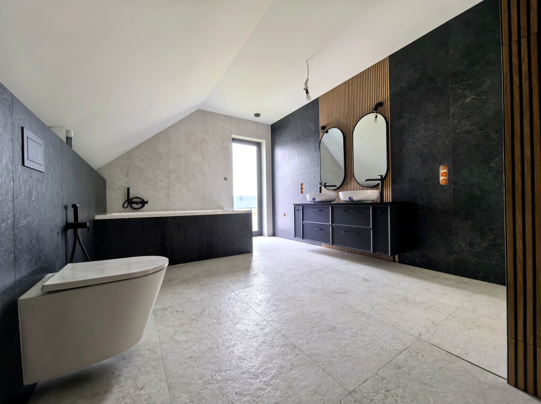 Nowoczesna łazienka na poddaszu z wanną, wiszącą toaletą i szafką z umywalkami. Ściany wykończone płytkami imitującymi kamień i drewnianymi lamelami.